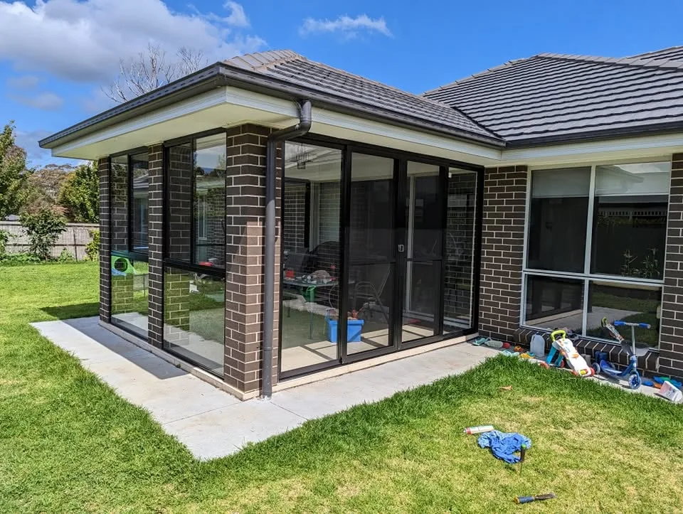 Brick home with a corner alfresco room enclosed by black aluminium sliding glass doors beside a green lawn